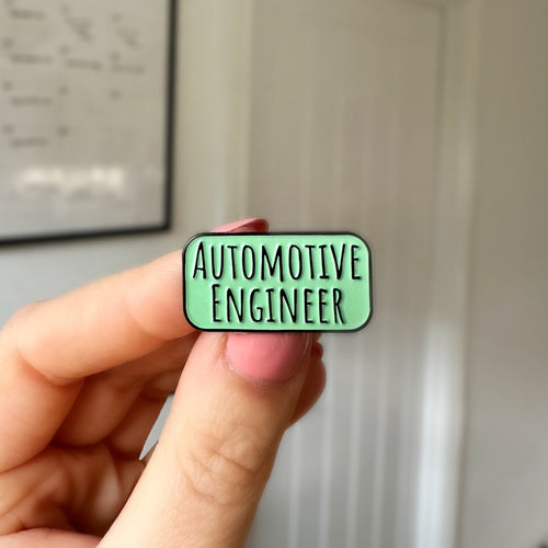 A hand holding a small rectangular enamel pin that reads 'AUTOMOTIVE ENGINEER' in black text on a light green background, with a blurred indoor room behind it.