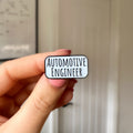 A hand holding a small enamel pin that reads 'AUTOMOTIVE ENGINEER' in black text on a white rounded rectangle with a thin black border, set against a blurred indoor background of a white door.