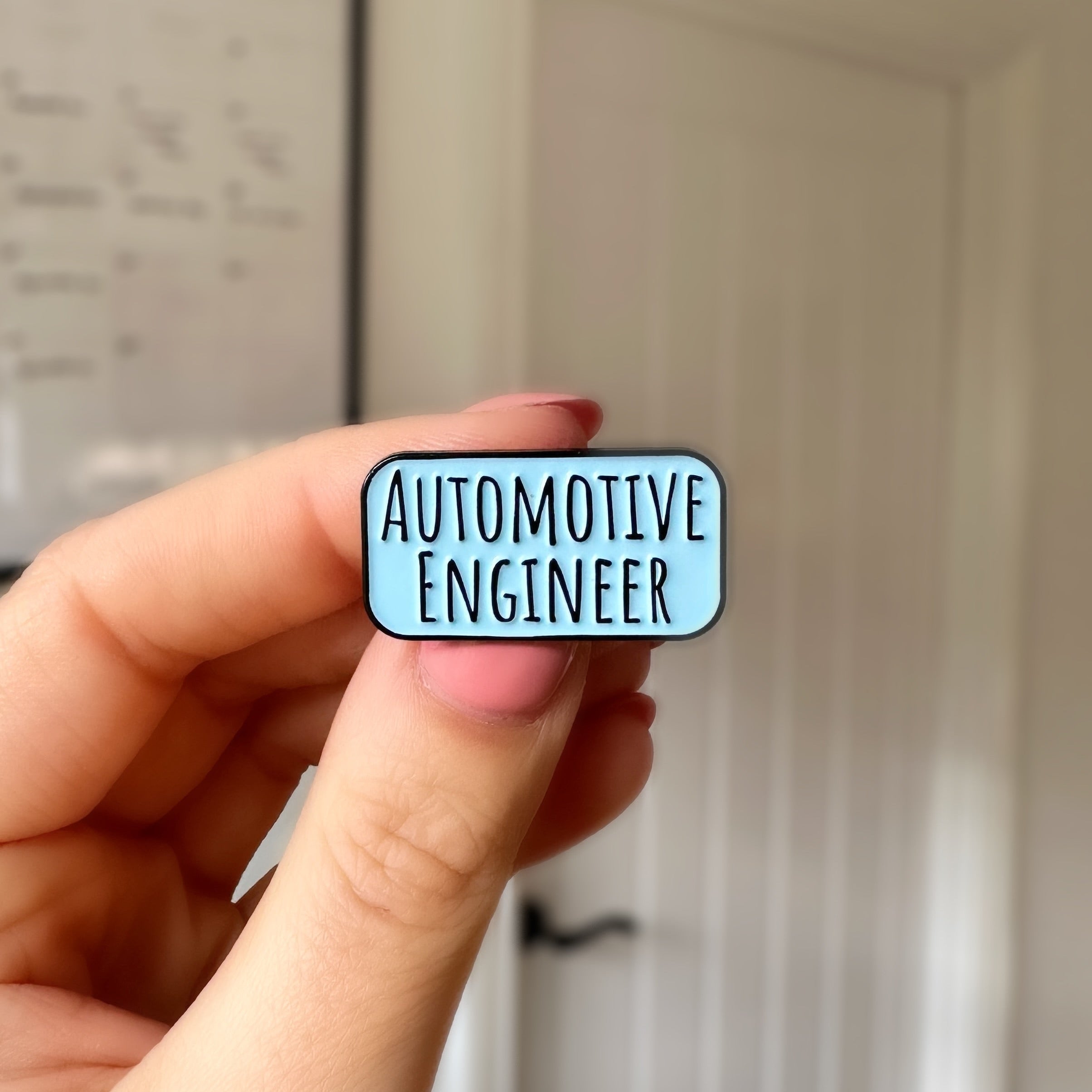 A hand holding a small enamel pin that reads 'AUTOMOTIVE ENGINEER' in black text on a light blue rounded rectangle background, with a blurred indoor room behind it.