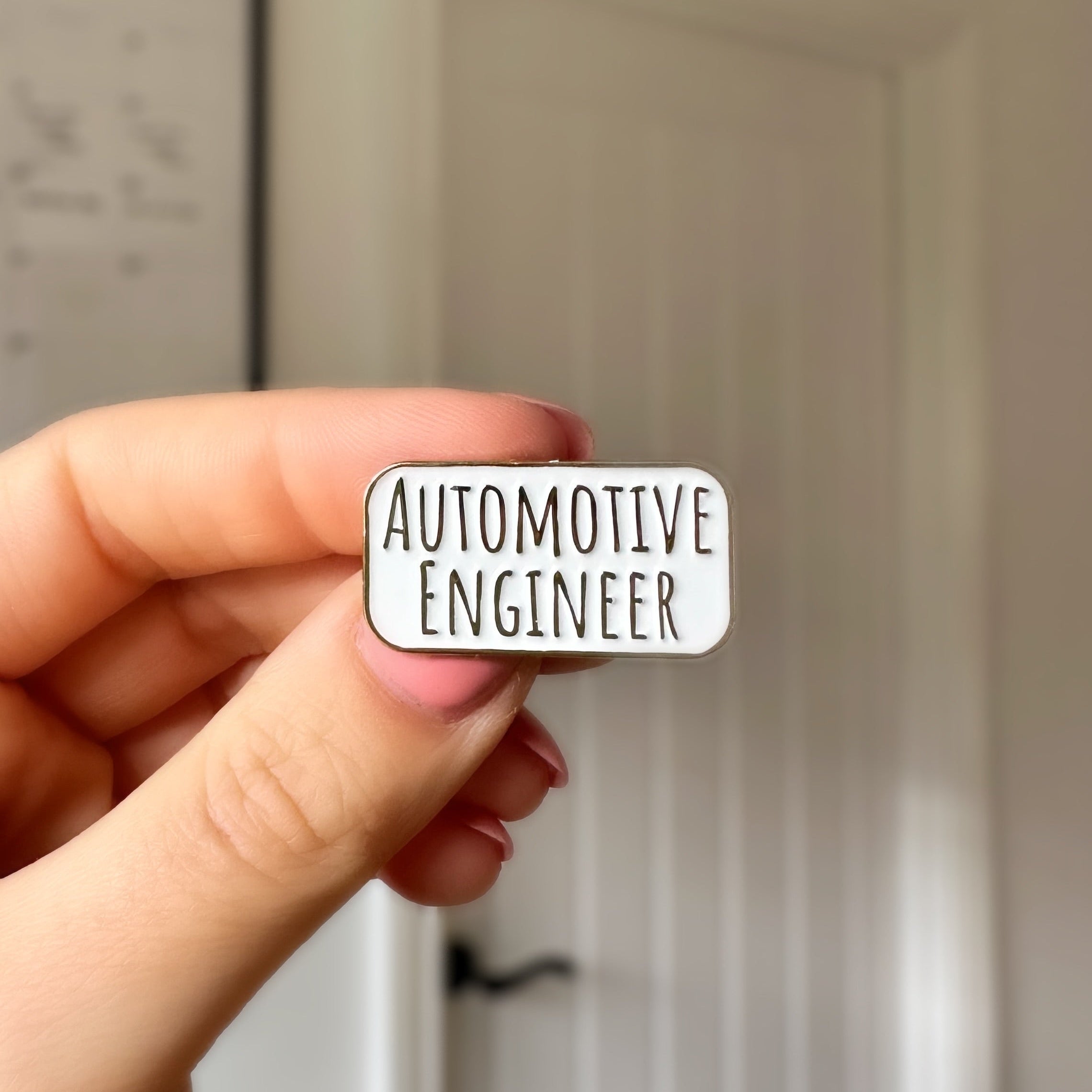 A hand holding a small white enamel pin with the words 'AUTOMOTIVE ENGINEER' in black lettering, set against a blurred indoor background of a white door.