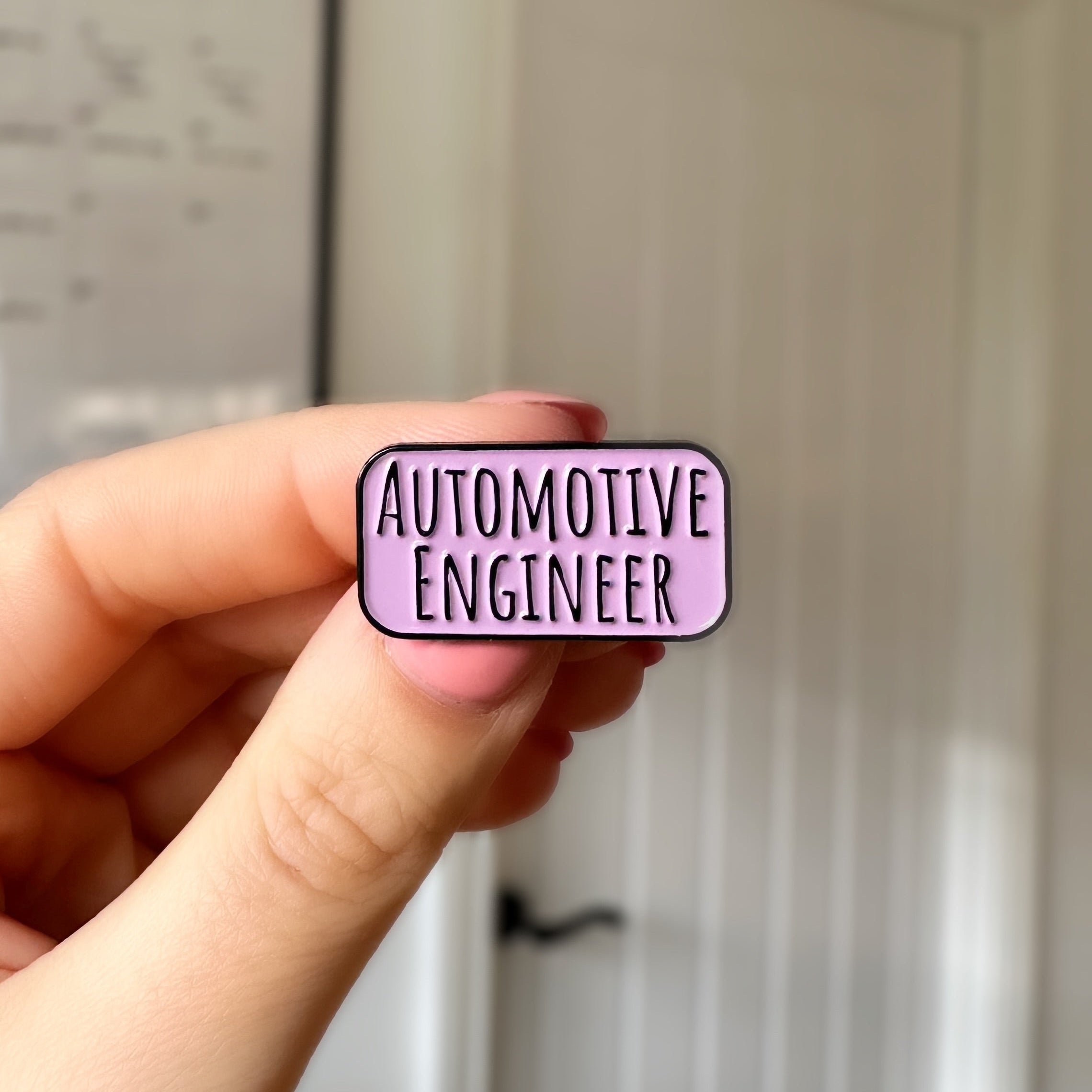 A hand holding a small rectangular enamel pin with rounded corners that reads 'AUTOMOTIVE ENGINEER' in black text on a light purple background, with a blurred indoor door in the background.